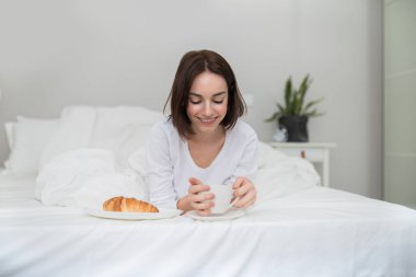 Breakfast in bed. Happy relaxed pretty young woman in white pajamas chilling in bed alone, holding tea mug, drinking coffee, eating pastry homemade croissant in bed, copy space