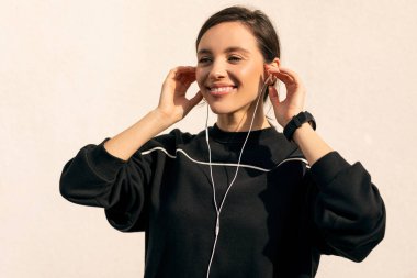 Cheerful caucasian millennial lady in sportswear and headphones enjoys fitness and jogging outdoors on white wall background, close up. Sports and music, body care and weight loss, active lifestyle