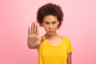 Serious confident young african american woman in yellow t-shirt make stop sign with hand isolated on pink background. Anti discrimination, fighting with harassment and domestic violence gesture