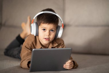 Serious caucasian little kid in headphones enjoys vacation, watching video, playing game on tablet lies on sofa, enjoy rest. Study at home remotely, childhood and video call, education, learning app