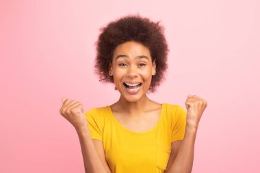 Cheerful excited young african american curly lady in yellow t-shirt with open mouth make victory and success gesture with hands, look at camera isolated on pink background, studio. Good luck emotion