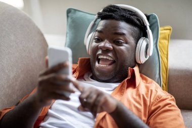 Closeup photo of emotional chubby millennial black guy in casual lying on couch with cell phone and wireless headphones, watching content on Internet, enjoying nice entertaining app, copy space