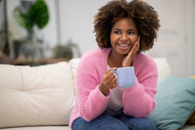 Happy curly millennial african american woman with teeth braces in homewear sitting on couch alone, drinking or coffee tea at home, looking at copy space and smiling