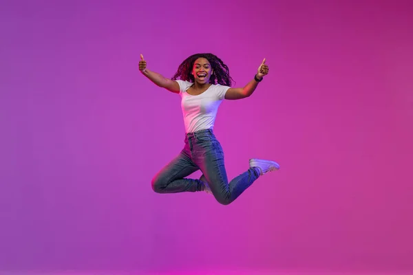 Cheerful Black Woman Jumping In Neon Light And Showing Thumbs Up At Camera, Positive Happy African American Lady Recommending Something And Having Fun Over Purple Background, Full-Length, Copy Space