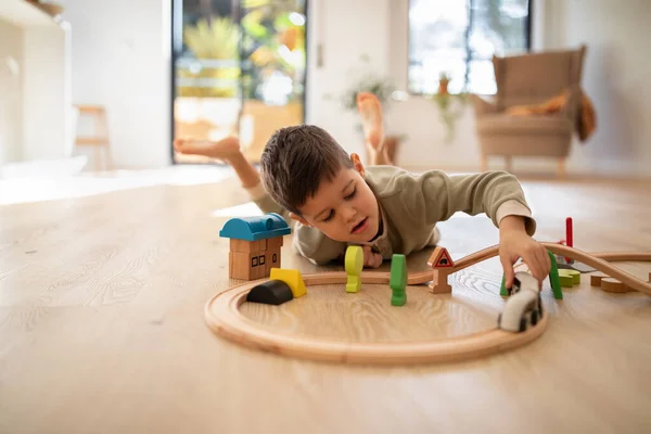 Busy cheerful caucasian little kid builds city with toys, plays road, train and cars, enjoy travel in kindergarten room interior. Fun alone, entertainment and education at home, fantasy and childhood