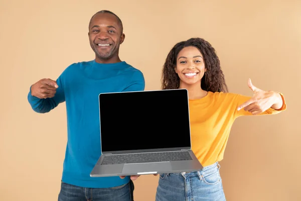 Excited black couple holding and showing laptop with black screen, presenting and pointing at gadget with blank space for mockup, peach studio background
