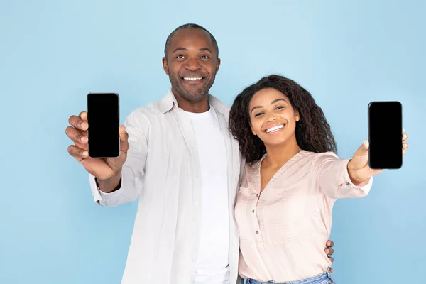 African american spouses showing modern smartphones with blank screens and smiling, recommending nice dating mobile application or website, mockup