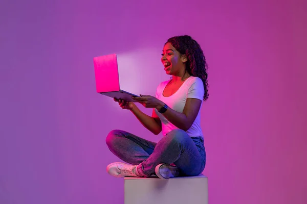 Excited Black Woman Looking At Glowing Laptop Screen While Sitting On Big Cube In Neon Light Over Purple Background, Cheerful Young African American Lady Enjoying Online Offer Or New Website
