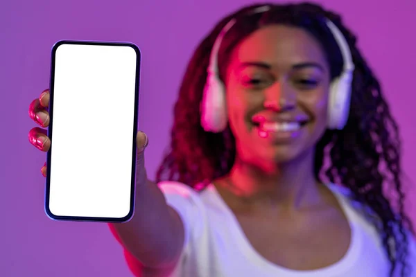 Cheerful Black Woman In Wireless Headphones Holding Blank Smartphone With White Screen In Hand While Standing In Neon Light Over Purple Backgtound, Happy Female Recommending Mobile App, Mockup