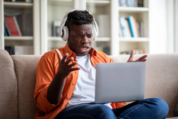 Telecommunication concept. Angry overweight african american young guy with stylish hairstyle having video conference while staying home, sitting on sofa, using modern laptop and wireless headphones