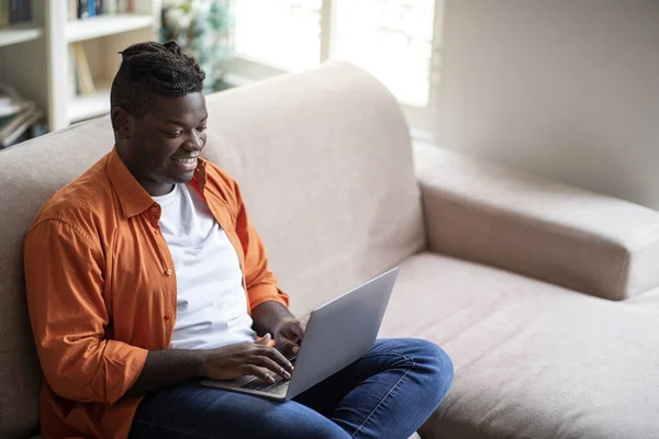 Remote job concept. High angle view of happy positive handsome chubby young black guy freelancer sitting on couch in living room, working from home, using laptop, copy space for ad