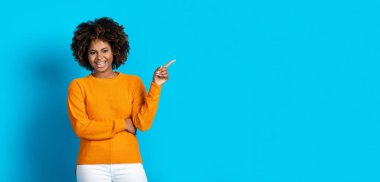 Cheery happy cool attractive curly millennial african american lady with teeth braces gesturing, pointing at copy space for advertisement on blue studio background, panorama