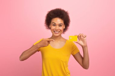 Cheerful young african american curly lady shopaholic in yellow t-shirt point finger at credit card, recommend sale isolated on pink background, studio. Finance and money for shopping, ad and offer