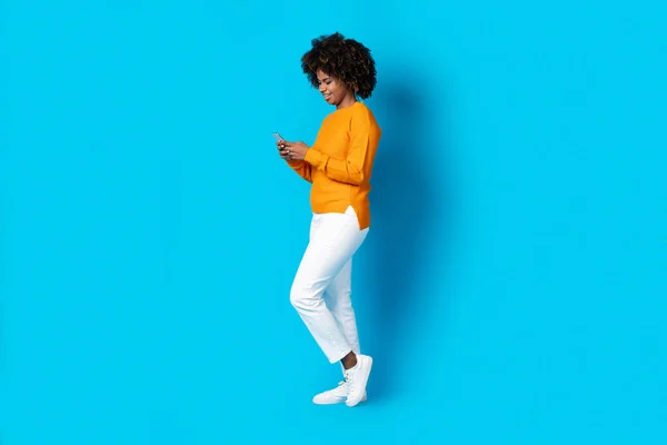 Dating mobile app. Happy cheerful millennial african american lady in stylish casual outfit with bushy hair and teeth braces chatting with guys, using cell phone on blue studio background, copy space