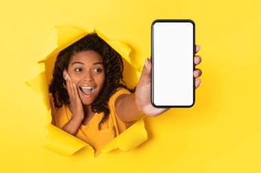 Surprised black woman showing cellphone with blank screen and stretching arm through hole in torn yellow paper background, advertising great offer, mockup
