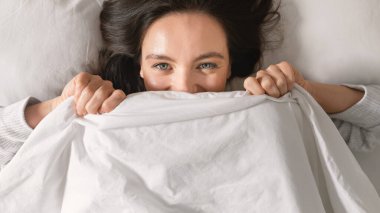 Cheerful caucasian millennial female lies on comfortable bed, enjoys softness, weekend, vacation and free time, has fun and covers face with blanket in bedroom interior. Good morning and rest at home