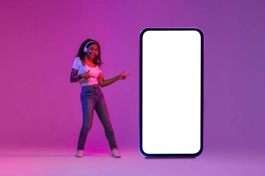 Check This. Cheerful Black Female Pointing At Big Blank Smartphone With White Screen While Standing In Neon Light Over Purple Gradient Background, Happy Lady Recommending New App, Collage, Mockup