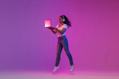 Positive African American Female Jumping With Laptop In Neon Lighting, Cheerful Black Lady Wearing Wireless Headphones Using Computer For Entertainment, Enjoying Modern Technologies, Copy Space