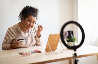 Beauty blog, blogging concept. Cheerful pretty curly young mixed race plump woman in casual outfit sitting at desk in front of mirror at home, recording video while applying makeup, using blogger set