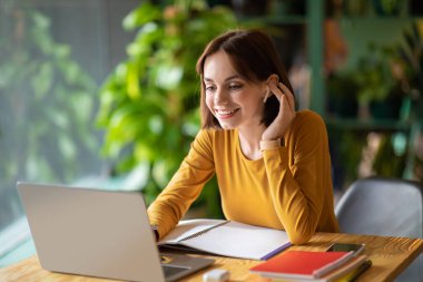 Cheerful beautiuful brunette young woman in smart casual entrepreneur attending online business meeting, woman sit at table in front of laptop, using earpods, taking notes, cafe interior, copy space