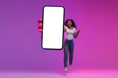 Joyful Black Lady Jumping With Big Blank Smartphone In Hand, Happy African American Woman Recommending New Mobile App Or Website, Having Fun In Neon Light Over Purple Background, Collage, Mockup