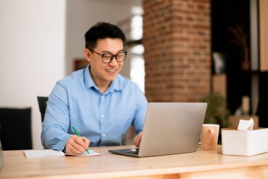 Happy asian man looking at laptop screen and writing in notebook, sitting at workplace at home office, selective focus. Male taking notes, watching webinar studying online