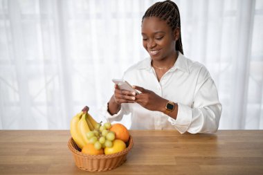Happy attractive young black woman in smart casual taking photo of bowl with fresh fruits on wooden table, using smartphone. Food blogger making content for blog about healthy diet, detox, copy space