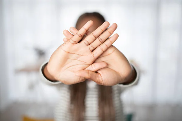 Unrecognizable long-haired black woman hiding behind her hands, showing both palms at camera, home interior, blurred background, copy space. Privacy, human rights concept