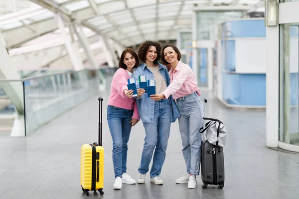 Three Happy Women With Passports And Tickets In Hands Posing At Airport Before Flight, Group Of Female Friends Standing With Suitcases At Terminal Hall, Enjoying Travelling Together, Copy Space