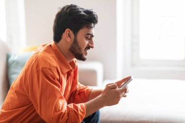 Happy attractive bearded young arab guy in orange shirt chilling at home, sitting on couch in cozy living room, using brand new smartphone, chatting with ladies on dating app, copy space, side view