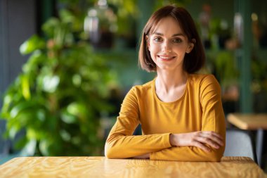Portrait of beautiful cheerful young brunette woman with nice haircut smiling at camera while chilling at cafe, coffee shop, sitting at table alone, copy space. Women in business, entrepreneurship