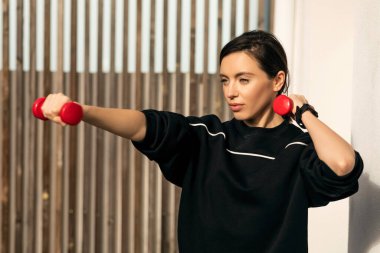 Serious caucasian millennial strong lady in sportswear doing exercises with dumbbells with hands on street, close up. Sports in city, fitness for weight loss, workout and active, body and health care