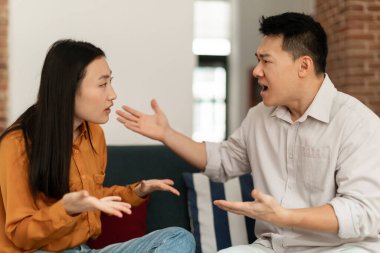 Angry asian spouses having quarrel experiencing crisis in relationship, sitting together on sofa at home. Middle aged man and yougn woman having conflict breaking up