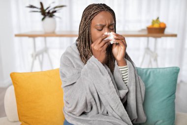 Ill unhappy millennial long-haired black lady sitting on couch covered in warm blanket at home, sneezing, using napkin, suffering from cold, flu or coronavirus, copy space, closeup