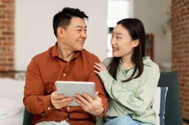 Happy asian lovers middle aged man and young woman sitting on sofa and using digital tablet, looking at each other and smiling, enjoying newest mobile application, home interior, copy space