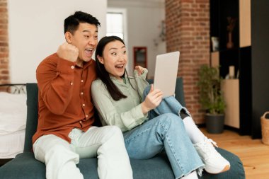 Overjoyed japanese couple sitting on couch with digital tablet, celebrating online win or business success at home, copy space. Asian middle aged man and his young wife enjoying big sale in web store