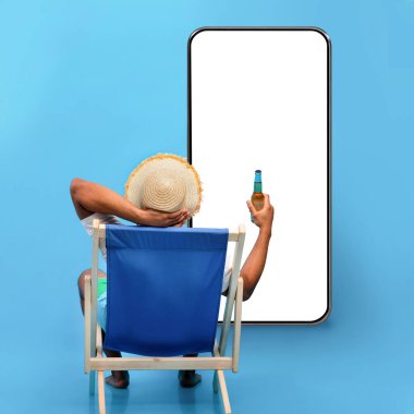 Back view of young black guy sitting in lounge chair with bottle of beer, sipping alcoholic beverage, looking at phone with blank screen on blue studio background, mockup for travelling offer, collage