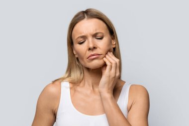 Closeup of sad middle aged attractive blonde lady touching her cheek, experience strong toothache, isolated on grey studio background, copy space, tooth pain concept