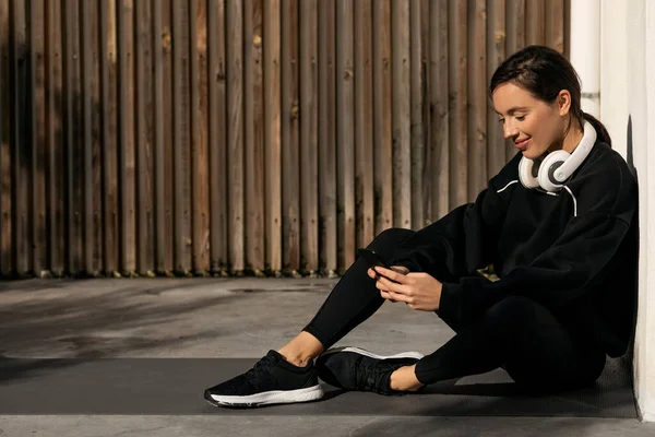 穿着运动服 头戴耳机 坐在地板上 在街上的智能手机上打字的高加索千禧年女士微笑着 运动博客 健身设备 运动应用 身体护理和减肥 — 图库照片