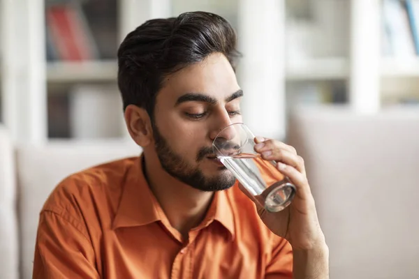 Healthy good-looking bearded millennial arab man in orange shirt sitting on couch, drinking fresh spring water at home, closeup, copy space. Hydration, healthy lifestyle concept