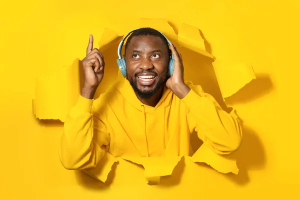 Excited black man enjoying music via wireless headphones and dancing in paper hole against yellow background. People lifestyle hobby concept