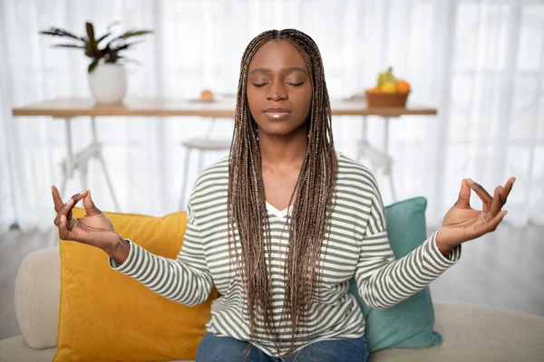 Stress relief. Relaxed peaceful pretty young black woman in casual outfit meditating on couch at home, sitting on sofa with closed eyes, hold hands in zen gesture, relaxing after bad day, copy space