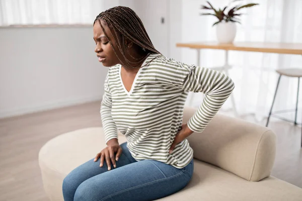 Upset young black long-haired woman in pain sitting on couch, touching her lower back, african american lady suffering from backache, muscle strain, period cramps, copy space