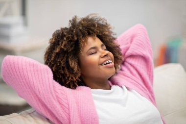 Closeup of happy joyful pretty young black woman in casual outfit chilling on couch at home, reclining on sofa with closed eyes and arms behind her head, dreaming about wealthy future, copy space