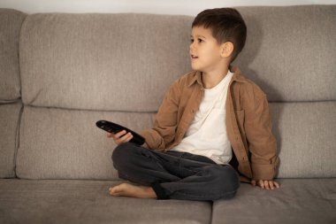 Serious cute european small kid uses remote control, switches channel on TV set, enjoy spare time alone, sits on sofa in living room, empty space. Education with device at home, cartoons and video