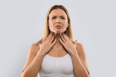 Unhappy middle aged blonde woman touching her neck, suffering from pain in throat, checking enlarged adenoids isolated on grey studio background, copy space. Sore throat concept