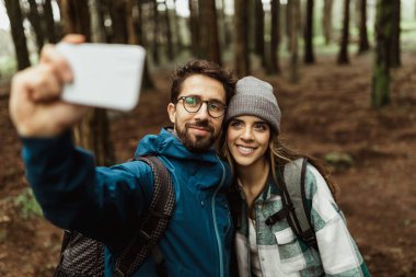 Smiling young european couple tourists in jackets with backpack walk in autumn forest, take selfie on smartphone, enjoy cold season outdoor. Lifestyle, app for travel, adventure and photo for blog
