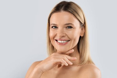 Portrait of cheerful attractive middle aged blonde woman smiling at camera, posing topless on grey studio background, demonstrating glowing perfect skin, copy space. Cosmetology concept