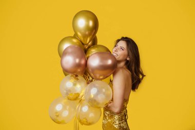 Festive concept. Excited woman with bunch of air balloons having holiday party over yellow studio background. Cheerful lady having fun on her birthday celebration