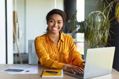 Portrait of happy black businesswoman working on laptop and smiling at camera, sitting at table in office interior, copy space. Cheerful woman sitting at workplace
